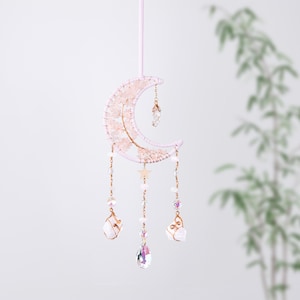 May include: A pink and gold wire-wrapped crescent moon with crystals and beads hanging from it. The moon is decorated with pink and white beads and a gold star. The crystals are clear and iridescent.