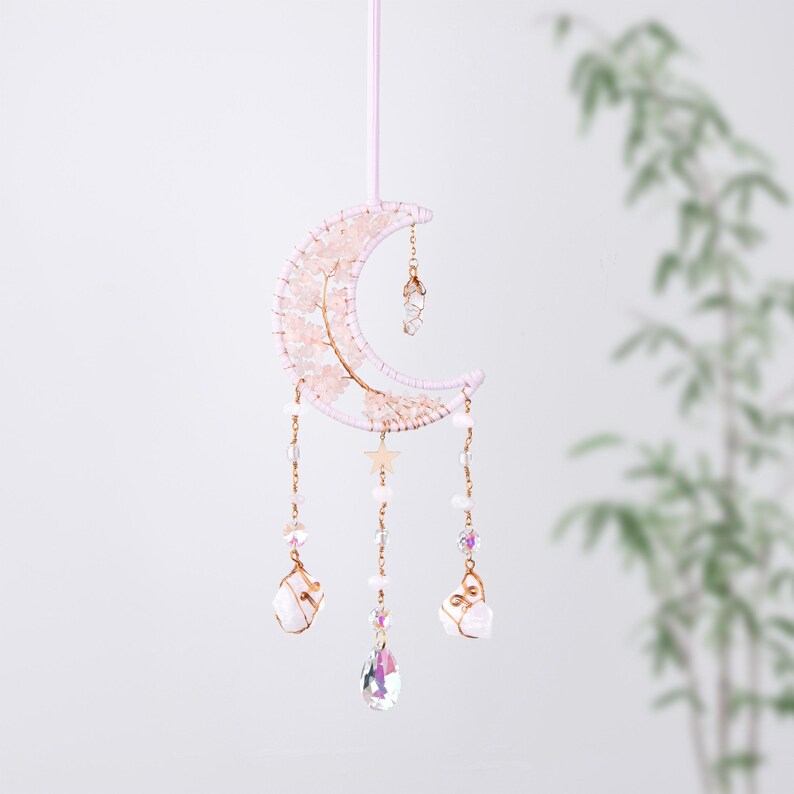 May include: A pink and gold wire-wrapped crescent moon with crystals and beads hanging from it. The moon is decorated with pink and white beads and a gold star. The crystals are clear and iridescent.