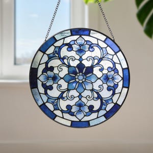 Op de afbeelding: Rond gebrandschilderd glazen raam paneel met een bloemenontwerp in blauw- en wittinten. Het paneel is omlijst met een donkerblauwe rand en wordt opgehangen aan een ketting. Het ontwerp toont gestileerde bloemen en wervelende patronen.