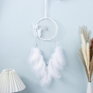Puede incluir: Un atrapasueños blanco con un marco circular, adornado con plumas blancas, una mariposa y cuentas colgantes. El atrapasueños está suspendido contra una pared azul claro, creando un acento decorativo.