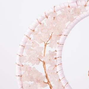 May include: A pink and gold wire tree with pink rose quartz crystals. The tree is wrapped around a white crescent moon shape.