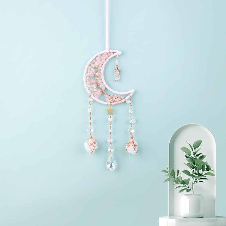 May include: A pink and gold moon-shaped dream catcher with crystal accents hanging from a white wall. The dream catcher has a star and small crystals hanging from it.