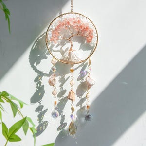 May include: A decorative dream catcher with a gold-toned metal ring. The centrepiece is a tree design crafted from pink and clear crystals. Dangling below are strands adorned with crystals and gemstones in diverse shapes and colours, creating a unique wall hanging.