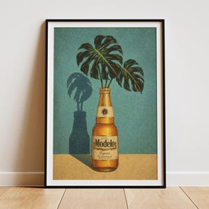 May include: A framed art print featuring a golden Modelo Especial beer bottle with two large green monstera leaves emerging from the top. The bottle sits on a tan surface against a textured teal background, casting a dark shadow to the left.