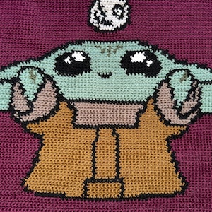 Puede incluir: Una manta de ganchillo con un personaje verde y marrón con ojos grandes. El personaje lleva un abrigo marrón y tiene un pequeño objeto blanco sobre su cabeza.