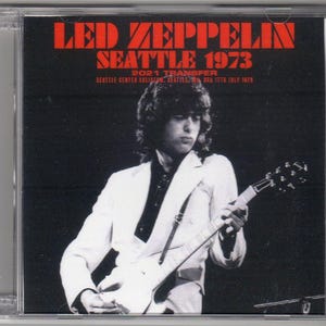 Könnte beinhalten: Ein Schwarzweißfoto eines Musikers, der eine weiße E-Gitarre spielt. Das Bild stammt von einem Konzert in Seattle, Washington im Jahr 1973. Der Text "LED ZEPPELIN SEATTLE 1973 2021 TRANSFER" ist auf dem Bild gedruckt.