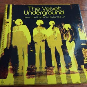 May include: A black and yellow album cover with the text "The Velvet Underground" and "Live at the Boston Tea Party '68 & '69". The cover features a black and white photo of four people standing in front of a yellow and black striped background.