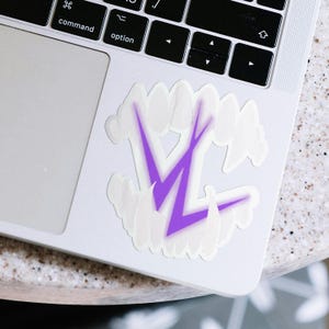 Könnte beinhalten: Ein weißer Aufkleber mit einem lila Blitzdesign, umgeben von weißen wolkenartigen Formen. Der Aufkleber befindet sich auf einem silbernen Laptop, wobei die Tastatur sichtbar ist. Das Design ist ein auffälliges, grafisches Element.