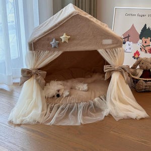 May include: A beige and white dog bed with a canopy and a small white dog sleeping inside. The bed has a ruffled edge and a bow on each side. The canopy is made of a sheer fabric and has two stars on the top.