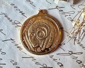 May include: A gold-colored pendant featuring the Aries zodiac sign, a ram's head with curled horns. The word "ARIETE" is inscribed around the top edge. The pendant is circular with a small loop at the top for a chain.