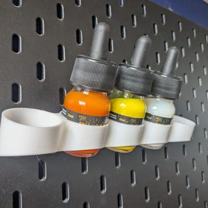 May include: Three small glass bottles with droppers, filled with orange, yellow, and white liquids. The bottles are held in a white plastic holder that is attached to a black pegboard.