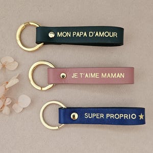 May include: Three leather keychains with gold rings. The first keychain is black with the text "MON PAPA D'AMOUR". The second keychain is pink with the text "JE T'AIME MAMAN". The third keychain is blue with the text "SUPER PROPRIO" and a gold star.