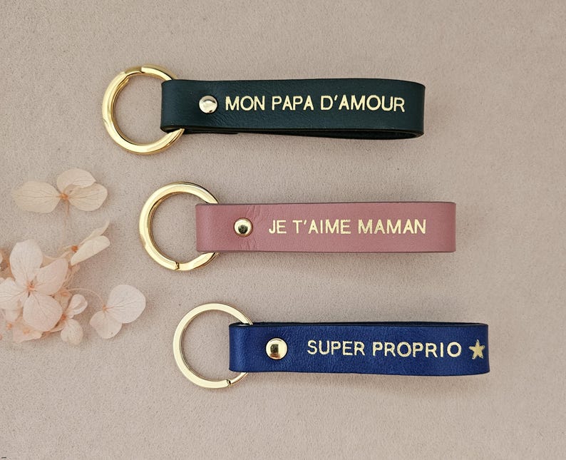 May include: Three leather keychains with gold rings. The first keychain is black with the text "MON PAPA D'AMOUR". The second keychain is pink with the text "JE T'AIME MAMAN". The third keychain is blue with the text "SUPER PROPRIO" and a gold star.