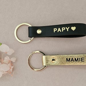 May include: Two leather keychains with gold key rings. The keychain on the left is black with the word "Papy" and a heart. The keychain on the right is gold with the word "Mamie" and a heart.