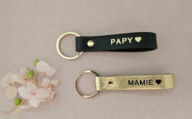 May include: Two leather keychains with gold key rings. The keychain on the left is black with the word "Papy" and a heart. The keychain on the right is gold with the word "Mamie" and a heart.
