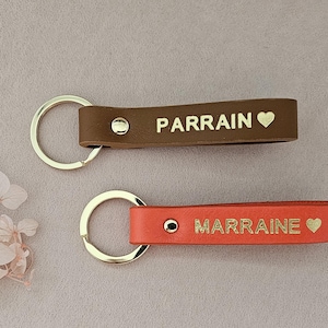 May include: Two leather keychains with gold lettering and a heart symbol. The top keychain is brown with the word "PARRAIN" and the bottom keychain is red with the word "MARRAINE".