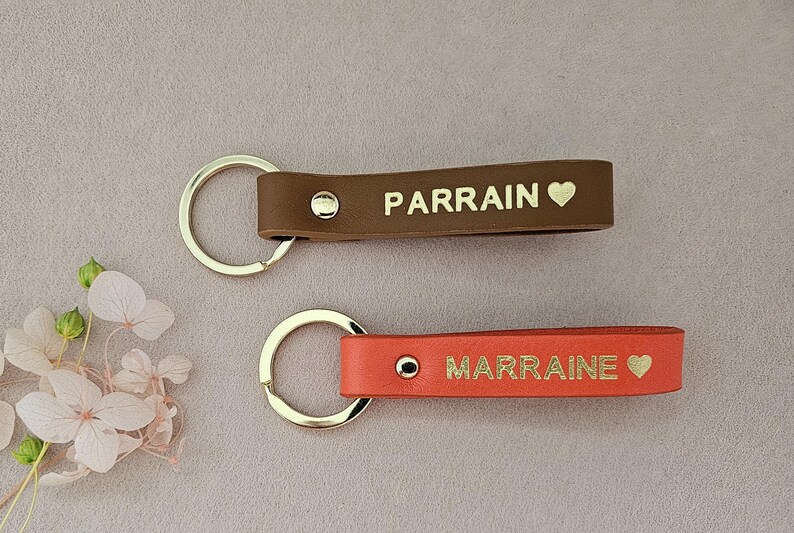 May include: Two leather keychains with gold lettering and a heart symbol. The top keychain is brown with the word "PARRAIN" and the bottom keychain is red with the word "MARRAINE".