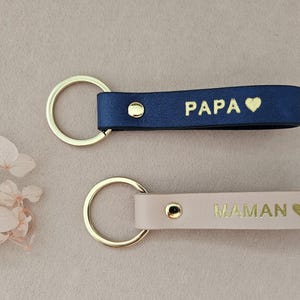 May include: Two leather keychains with gold rings. One is navy blue with the word "PAPA" and a heart. The other is beige with the word "MAMAN" and a heart.