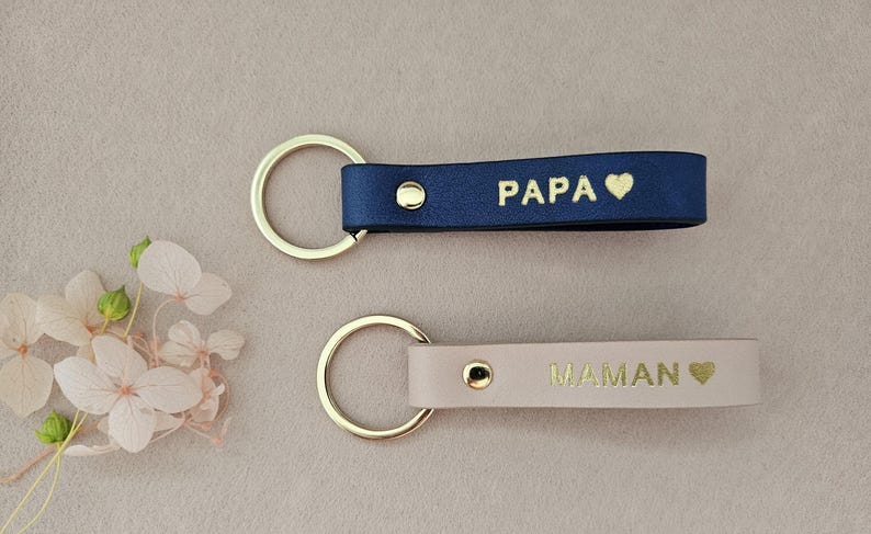 May include: Two leather keychains with gold rings. One is navy blue with the word "PAPA" and a heart. The other is beige with the word "MAMAN" and a heart.
