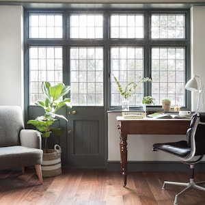 May include: A home office featuring a desk, chair, and a grey armchair. The desk is made of wood and has a white lamp on it. The chair is black with a silver frame. The armchair is grey with wooden legs. The room has large windows with black frames and a white radiator on the wall.