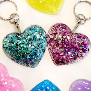 May include: Two heart-shaped keychains with glitter embedded in clear resin. One keychain is teal and silver glitter, the other is pink and purple glitter.