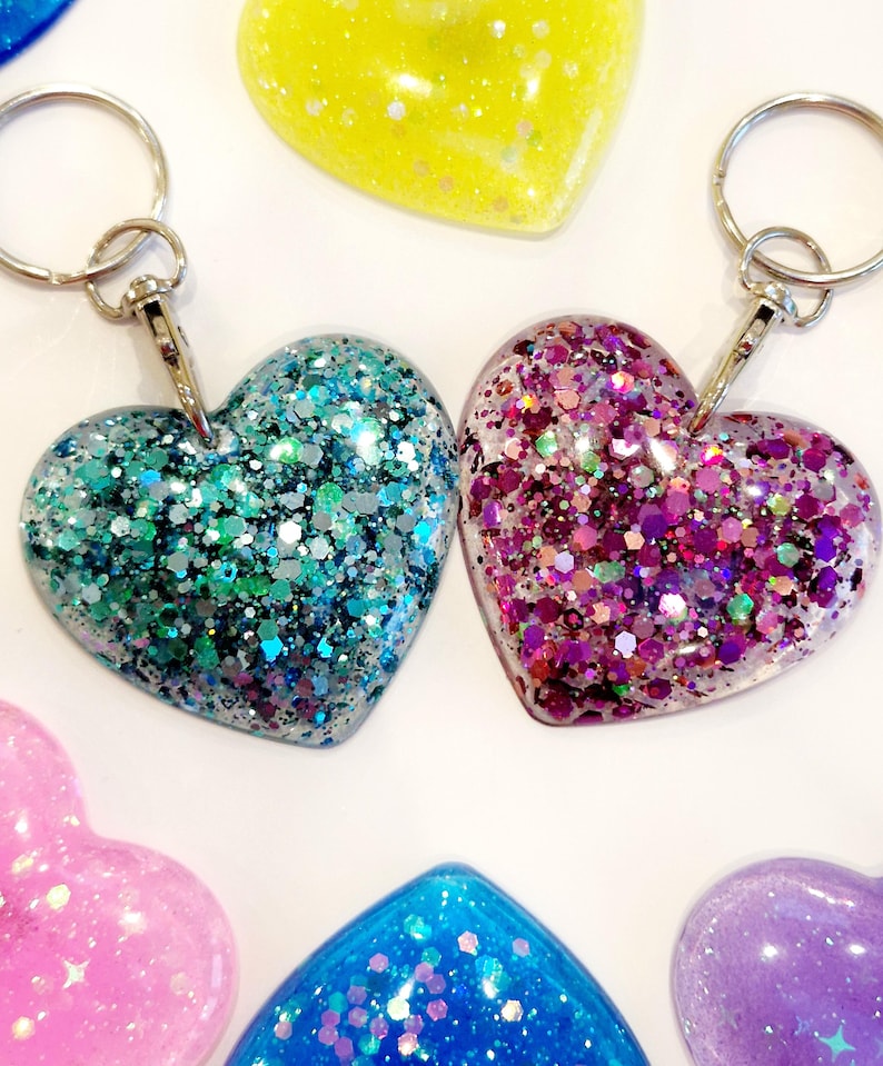 May include: Two heart-shaped keychains with glitter embedded in clear resin. One keychain is teal and silver glitter, the other is pink and purple glitter.