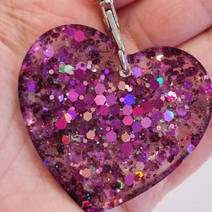 May include: A heart-shaped keychain made of clear resin filled with iridescent pink, purple, and green glitter. The keychain has a silver metal clasp.