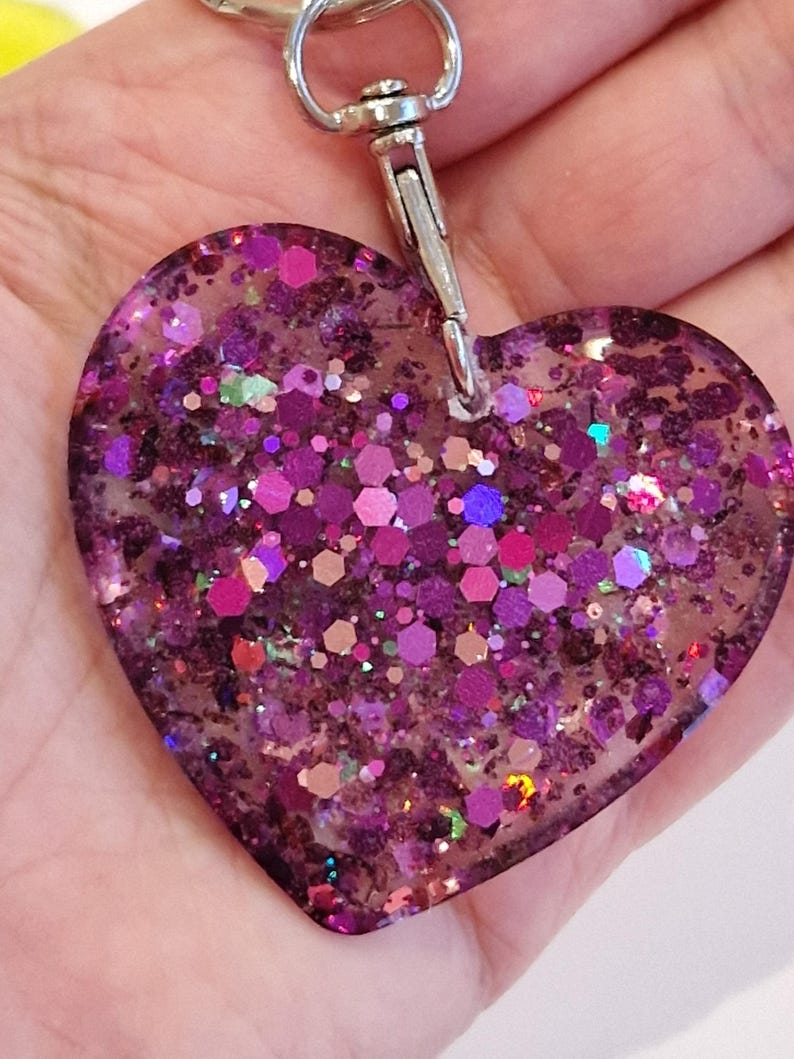 May include: A heart-shaped keychain made of clear resin filled with iridescent pink, purple, and green glitter. The keychain has a silver metal clasp.