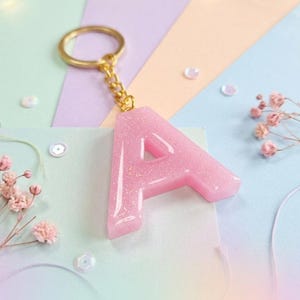 May include: A pink glitter resin letter "A" keychain with a gold-colored chain and keyring. The letter is set against a pastel background with decorative elements.
