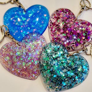 May include: Four heart-shaped keychains with glitter embedded in clear resin. The keychains are in shades of blue, pink, green, and purple.