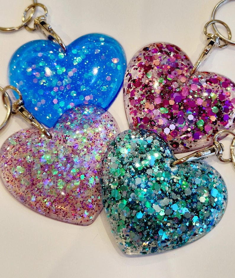 May include: Four heart-shaped keychains with glitter embedded in clear resin. The keychains are in shades of blue, pink, green, and purple.