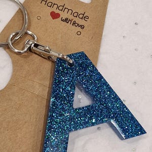 May include: A blue glitter letter A keyring with a silver ring and clasp. The keyring is attached to a brown card with the text "Handmade with love".