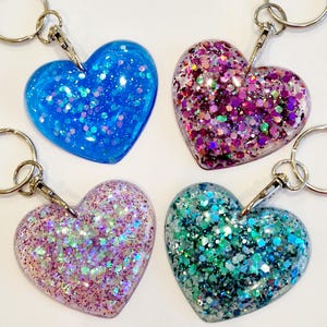 May include: Four heart-shaped keychains with glitter embedded in clear resin. The keychains are blue, pink, purple, and green.
