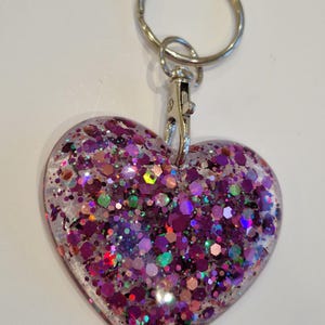 May include: A heart-shaped keychain with a silver key ring and clasp. The heart is filled with iridescent glitter in shades of pink, purple, green, and gold.