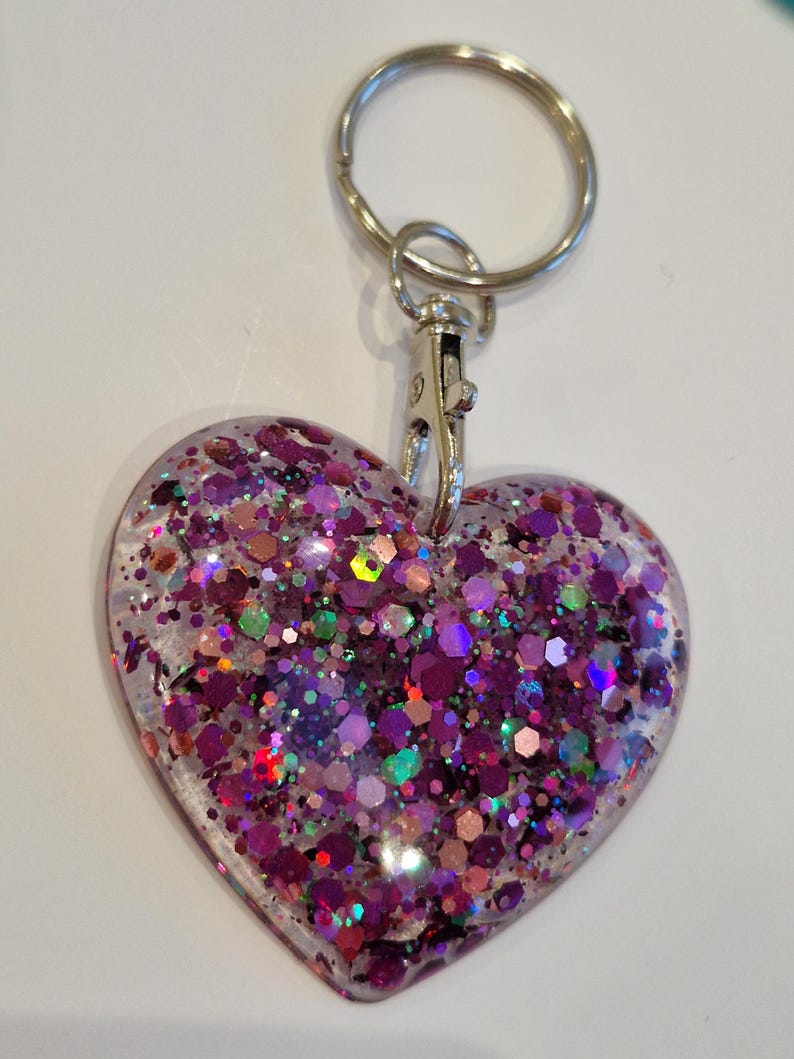 May include: A heart-shaped keychain with a silver key ring and clasp. The heart is filled with iridescent glitter in shades of pink, purple, green, and gold.