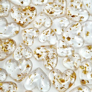 May include: A collection of clear, heart-shaped embellishments with gold leaf flakes scattered throughout. The hearts vary in size and are arranged closely together, creating a textured, romantic aesthetic. The gold leaf adds a touch of luxury to the transparent hearts.