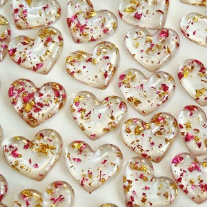 May include: A collection of small, heart-shaped resin charms featuring a clear, translucent base embedded with scattered pieces of metallic gold foil and vibrant pink flakes, arranged in a repeating pattern across the frame.