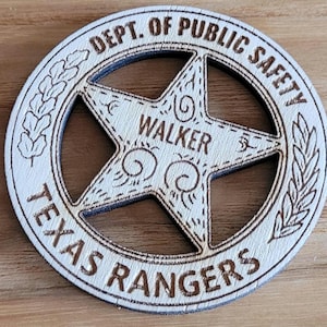 Peut inclure: Un badge circulaire en bois avec une étoile à cinq branches découpée au centre. Le texte DEPT. OF PUBLIC SAFETY et TEXAS RANGERS est gravé sur le bord, avec le nom WALKER gravé dans l'étoile centrale, entouré de motifs en volutes.