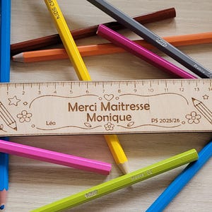 Puede incluir: Una regla de madera con texto grabado e ilustraciones de lápices, rodeada de lápices de colores. La regla dice "Merci Maitresse Monique" y "PS 2025/26". La regla mide aproximadamente 15 cm de largo.