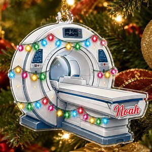 May include: A flat, circular ornament shaped like an MRI machine, featuring a white and blue scanner with a patient table. The machine is decorated with a string of colorful, glowing Christmas lights and displays the name Noah in red script.