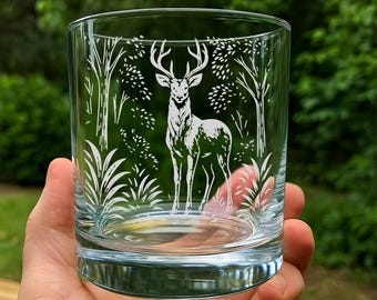 May include: A clear glass tumbler with a detailed white etched design of a deer standing in a forest scene. The design includes trees, foliage, and the deer's antlers. The glass is held in a hand against a blurred green background.