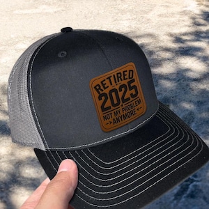 May include: A black and gray trucker hat with a brown leather patch that says "Retired 2025 Not My Problem Anymore".