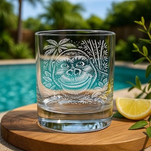 May include: A clear glass tumbler with a smiling gorilla design etched in white and teal. The design includes palm trees and bamboo. The glass sits on a wooden surface with a lemon wedge nearby, with a pool in the background.