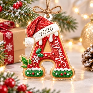 May include: A clear acrylic Christmas ornament shaped like the letter A, decorated with red and green icing details, holly leaves, and a white snowflake. It features a red Santa hat, the year 2026, and the name Amelia in white script lettering.