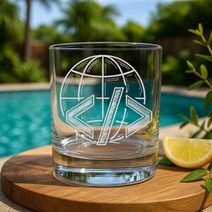 May include: A clear, lowball glass with a white etched design featuring a globe and code symbols. The glass sits on a wooden surface with a lemon wedge and greenery in the background. The glass is perfect for serving cocktails or other beverages.