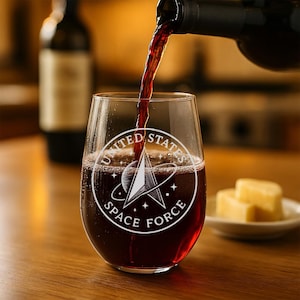 May include: A stemless wine glass being filled with red wine. The glass has a white graphic of the United States Space Force logo. A bottle of wine is in the background, and a small plate of cheese is to the right.