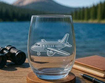 May include: A clear stemless wine glass with an etched airplane design. The glass sits on a wooden coaster. A pair of binoculars, a book, and a pen are also visible in the image. The background features a lake and a mountain.