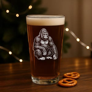 May include: A pint glass filled with beer, featuring a detailed white illustration of a gorilla. The glass sits on a wooden surface with two pretzels. The beer has a foamy head.
