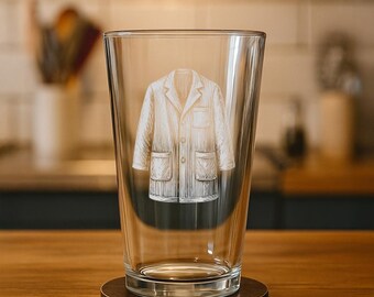 May include: A clear glass pint glass with a detailed etching of a lab coat. The glass sits on a round, dark brown coaster. The background is blurred, showing a kitchen setting with wooden surfaces and white tile.