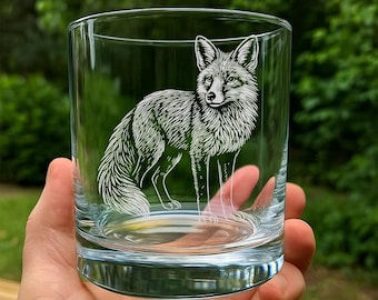 May include: A clear glass tumbler with a detailed white etched design of a fox. The fox is depicted in a standing pose, with its fur and features intricately rendered. The glass is held in a hand, with a blurred green background.